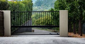 Black metal wrought iron driveway property entrance gates set in brick fence, concrete path, garden trees