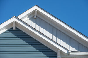 Double gable, with white decorative corbel, bracket, brace on a triangle gable roof, white soffit and fascia, gray vertical and blue horizontal vinyl lap siding with blue sky background